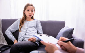 Young woman on couch in therapy session. Hands in foreground taking notes with clipboard and pen.